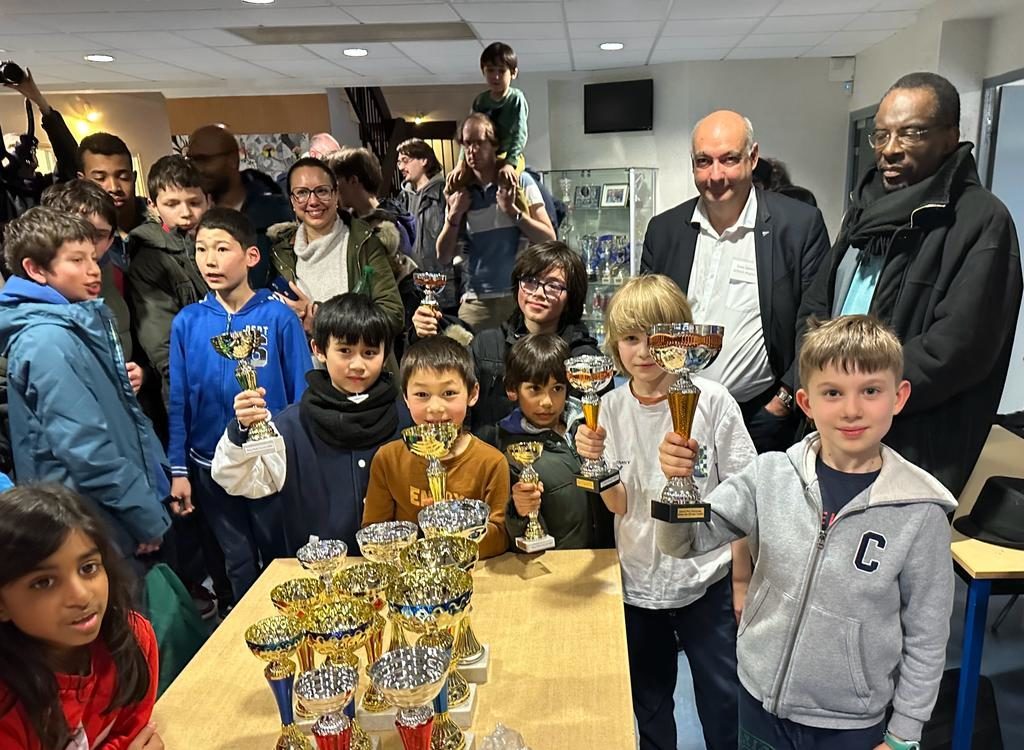 Groupe d'enfants brandissant leurs coupes devant la table des trophées, entourés des organisateurs.