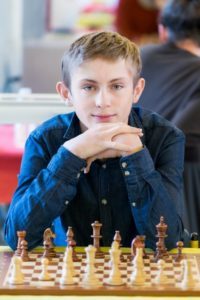 Un jeune joueur souriant, mains sous le menton, pose derrière son échiquier au tournoi de Rouen.